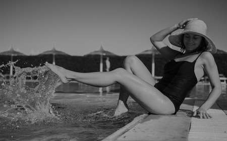 Seductive girl in a hat wets her feet while sitting on the edge of the pool. Vacation and recreation concept. Black and white photo.の写真素材