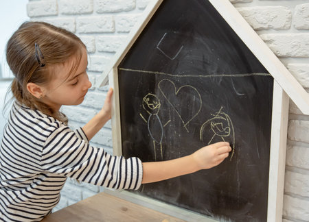 A little girl draws a chalk drawing on a blackboard in the shape of a house.の写真素材