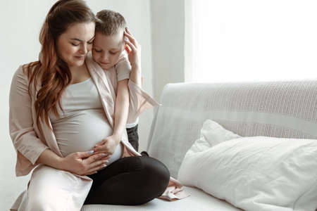 A young pregnant woman is sitting on the couch with her little son in an embrace.の写真素材