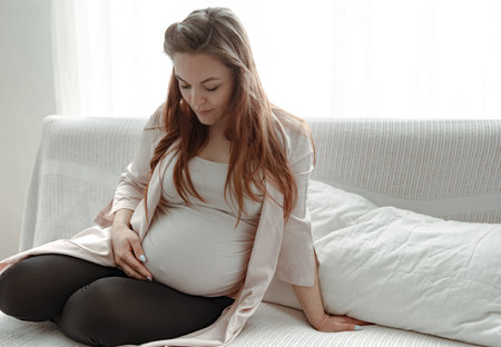 A young pregnant woman is resting on a sofa in a bright, cozy room.