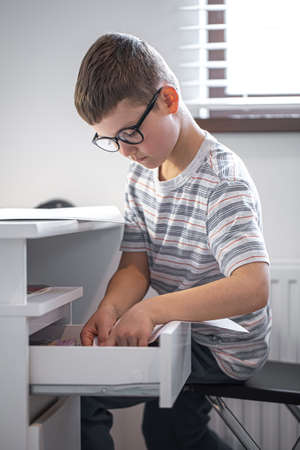 Little boy with glasses sitting at his desk in front of a laptop looking for something in a drawer.の写真素材