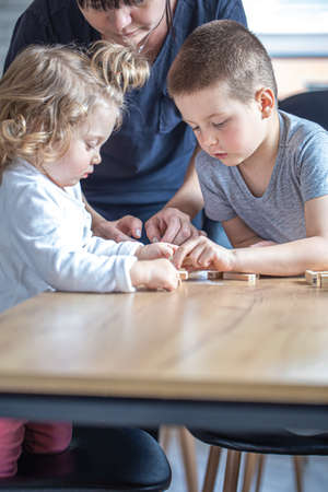 Little children play board game with wooden cubes at home in the kitchen with mom.の写真素材