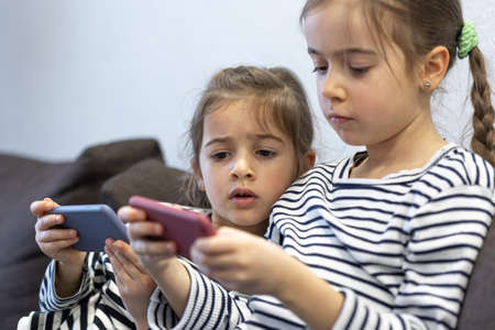 Cute little sisters use phones while sitting on the couch at home.の写真素材