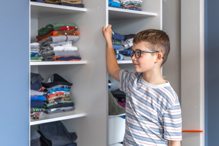 A cute boy with glasses stands near a wardrobe and thinks about what to wear.の写真素材