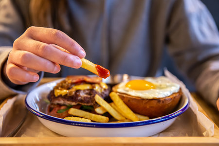 A girl holds fries in ketchup in her hand on a blurred background with a burger.