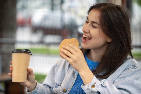 Attractive girl in casual style eats a burger with coffee sitting on the summer terrace.