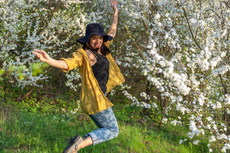 An attractive girl in a hat among blooming trees enjoys the smell of spring flowers.