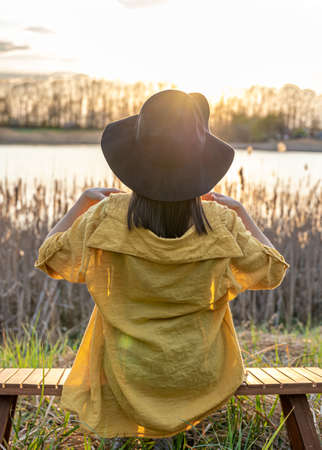 A girl in a hat and in casual style sits on a bench near the lake at sunset.の写真素材