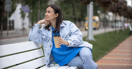Street photo of an attractive young woman sitting on a bench with a coffee in her hand.の写真素材