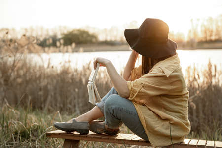 Stylish girl in a hat reads a book sitting by the river at sunset view from the back.の写真素材