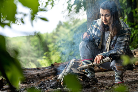 An attractive young woman makes a fire to keep warm in the forest.
