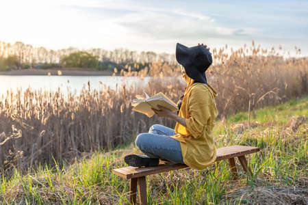 Attractive girl in a hat reads a book in nature at sunset.の写真素材