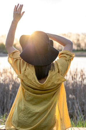 A girl in a hat and in casual style sits on a bench near the lake at sunset.