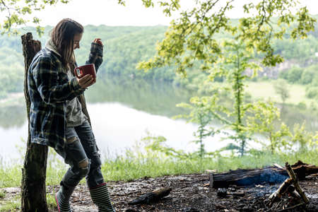 A young woman in the forest near the river warms up by the fire and drinks a hot drink.の写真素材