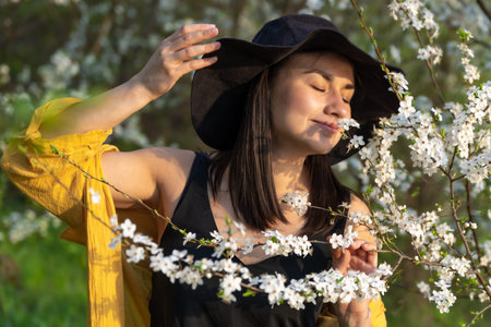 An attractive girl in a hat among blooming trees enjoys the smell of spring flowers.