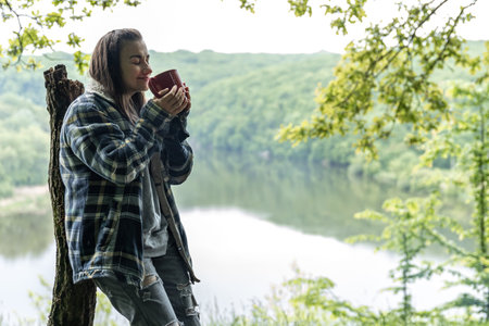 A young woman in the forest near the river warms up by the fire and drinks a hot drink.