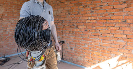 An electrician in a hard hat looks at the wall while holding an electric cable.の写真素材
