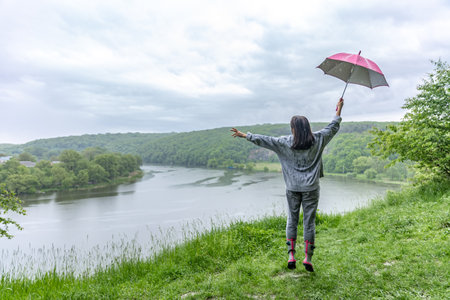 Back view of a girl under an umbrella jumping near a lake in a mountainous area in rainy weather.の写真素材