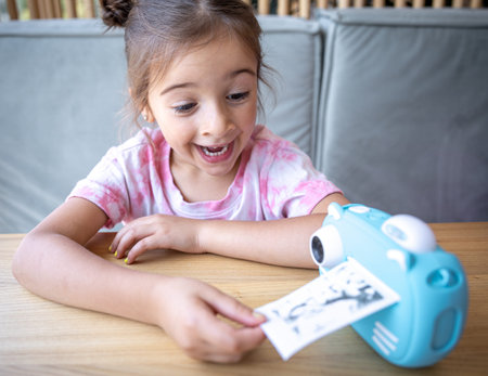A cute little girl looks at her children's blue toy camera for instant photo printing.