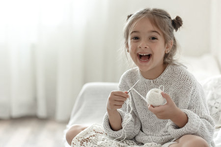 Happy little girl sitting on the sofa and learning to knit, home leisure concept.