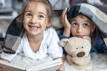 Little sisters are reading a book with a teddy bear lying on the floor in the room.