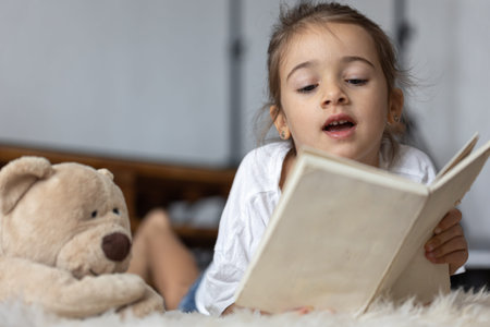 Cute little girl at home, lying on the floor with her favorite toy and reads book.の写真素材