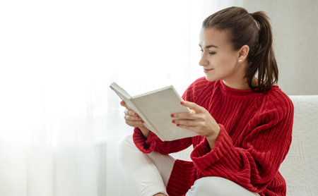 Young attractive woman in a cozy red sweater is reading a book while sitting on the couch at home.