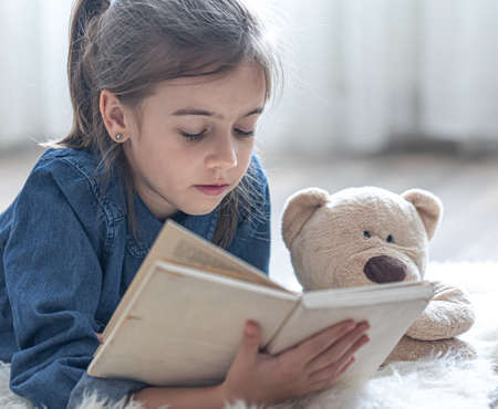 Pretty little girl at home, lying on the floor with her favorite toy and reads book.の写真素材