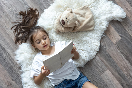 The child reads a book lying on a cozy rug at home with his favorite toy teddy bear.
