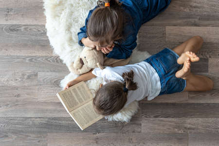 Little sisters are reading a book with a teddy bear lying on the floor in the room top view.の写真素材