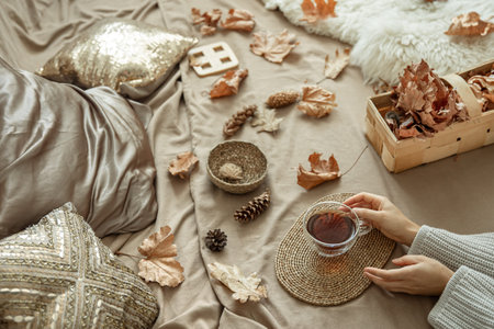 Home autumn composition with female hands and a cup of tea in bed, among pine cones and autumn...
