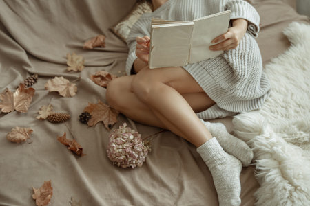 A girl in a cozy knitted sweater and warm socks lies in bed with a book among the autumn leaves.