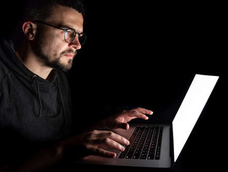 A man in glasses looks seriously at a laptop screen, works at a computer at night in the dark.の写真素材