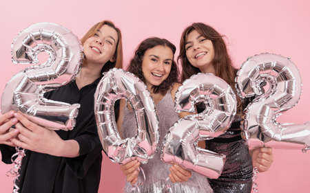 A company of happy girlfriends on a pink background with silver foil balloons in the form of the numbers 2022, celebrating the new year.の写真素材
