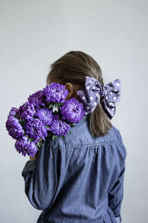 Little girl with a beautiful bow on her hair holds a bouquet of blue chrysanthemums, back view.の写真素材
