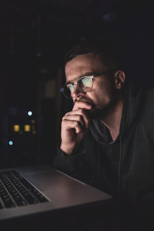 A man with glasses in the dark looks at the computer screen, copy space.