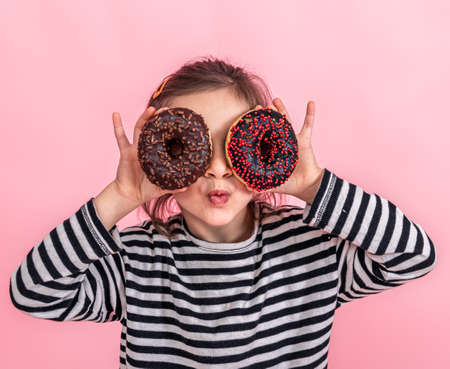 Portrait of a little smiling girl brunette with and two appetizing donuts in her hands, closes her eyes with donuts, on a pink background.の写真素材