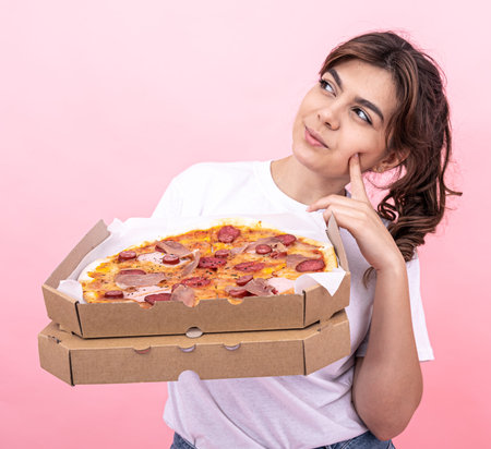 Cute pensive girl with a box of pizza for delivery on a pink background, copy space.の写真素材