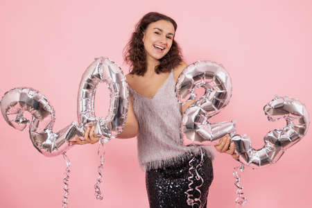 Beautiful brunette girl with curly hair and festive clothes posing on a pink background with confetti and holding silver balloons from the numbers 2023.の写真素材