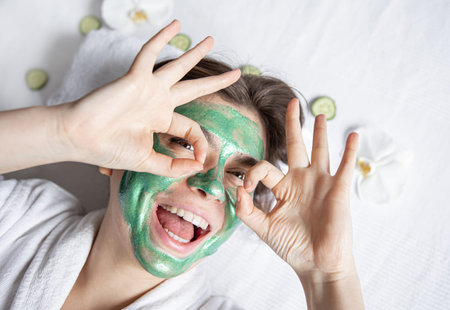 Funny young woman with a green cosmetic mask on her face is resting while lying in the spa salon,...