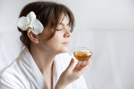A young woman in a white robe with an orchid flower in her hair is enjoying tea, the concept of spa...