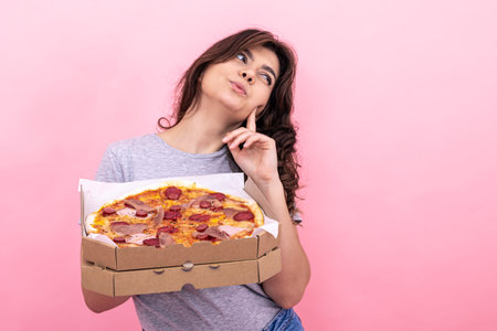 Cute pensive girl with a box of pizza for delivery on a pink background, copy space.の写真素材