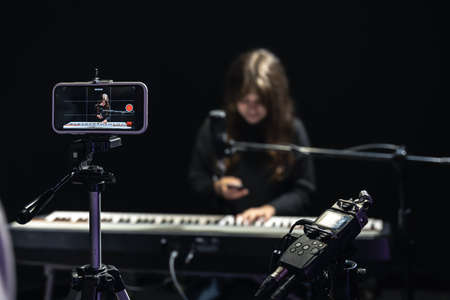 Girl musician recording video on smartphone standing on tripod, using professional microphone, blogger or music teacher shooting course in studio, sitting at the piano.の写真素材
