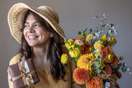 Cheerful young woman in a yellow dress and hat with a bouquet of bright chrysanthemums and a gift on...