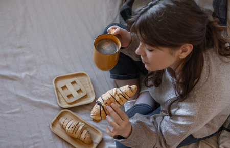Young woman drinking coffee with a croissant in bed, top view.