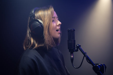 Cute young woman in headphones sings into a microphone in a dark room.