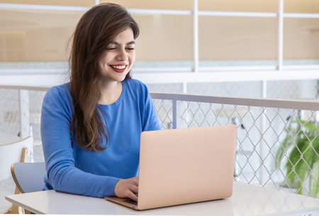 Smiling young woman in a blue sweater works at a laptop in a cafe.