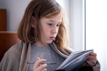 Serious little girl draws in a notebook, wrapped in a blanket.の写真素材