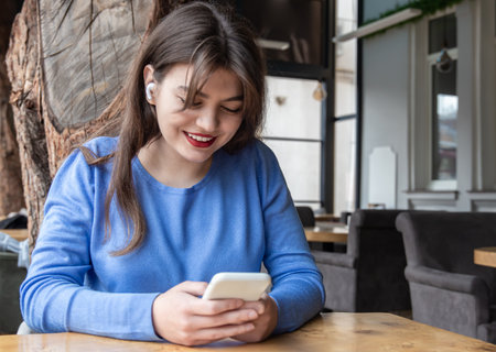 A young woman in wireless headphones uses the phone while sitting in a cafe.の写真素材