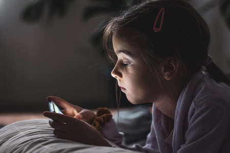 Little girl uses a smartphone lying on the pillow at home.の写真素材